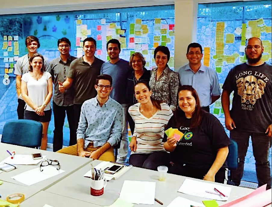 Um grupo de treze pessoas posa para uma foto em um ambiente de escritório, com post-its coloridos na parede atrás deles. Alguns estão sentados, enquanto outros estão de pé. Eles estão sorrindo e parecem estar em um ambiente colaborativo ou de reunião.